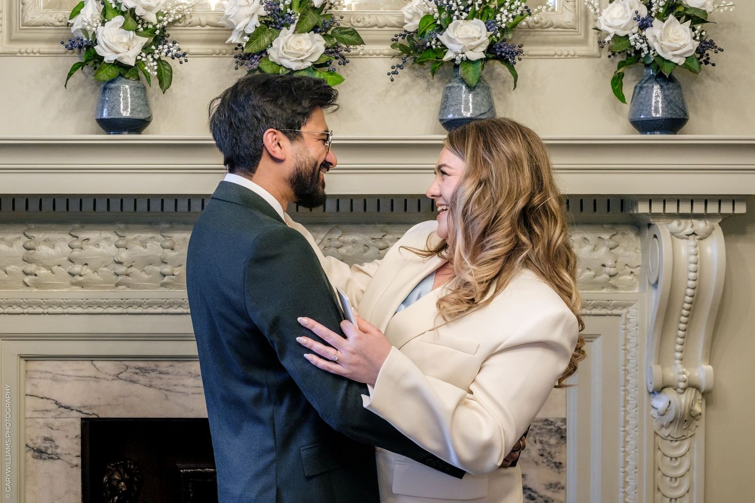 Couple sharing a romantic moment by an elegant fireplace adorned with white floral arrangements.