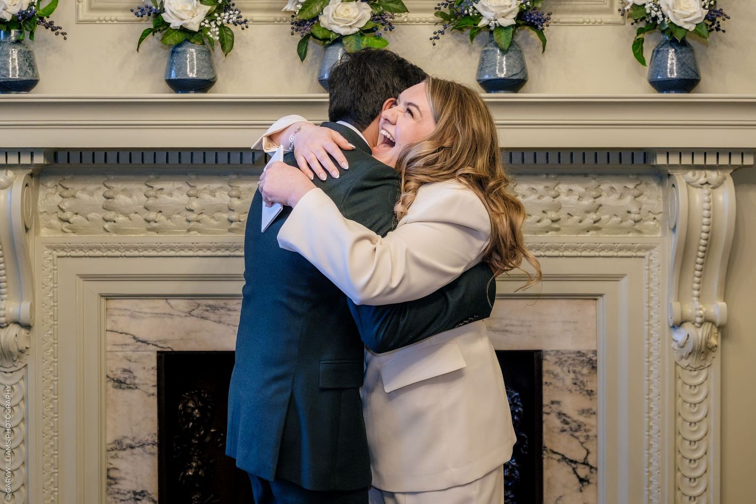 Couple embracing joyfully in front of an elegant fireplace with floral decor, dressed in formal wedding attire.
