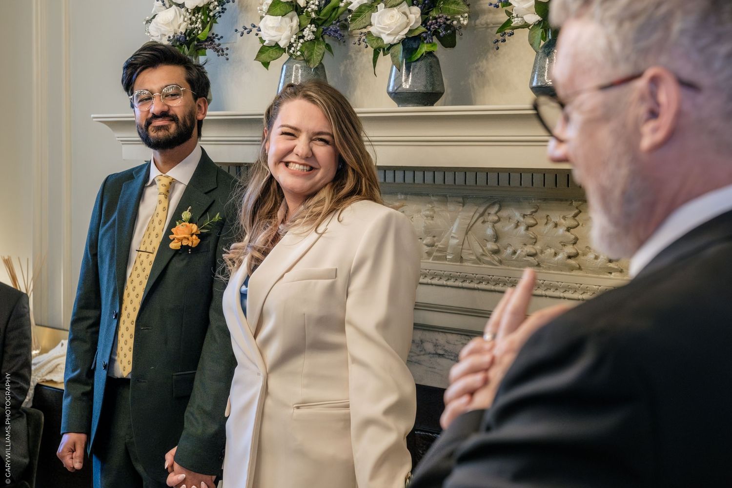 Bride in cream blazer smiles during wedding ceremony beside groom in teal suit with yellow floral tie.