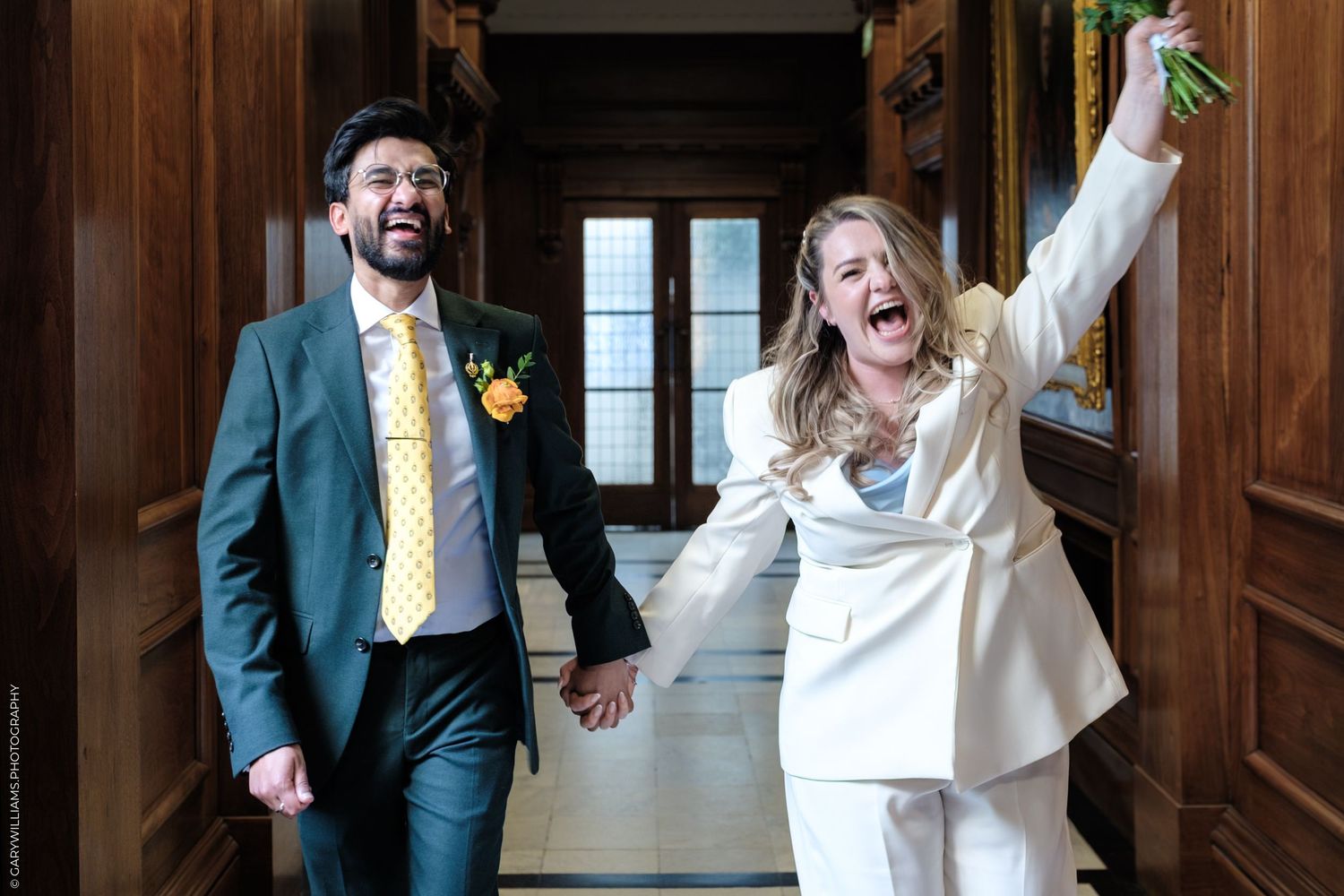 Joyful newlyweds holding hands, groom in teal suit with yellow tie, bride in white pantsuit raising bouquet.