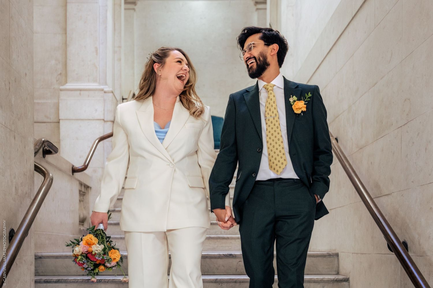 Happy newlyweds walk down stairs holding hands, bride in white suit with bouquet, groom in dark suit with yellow tie.