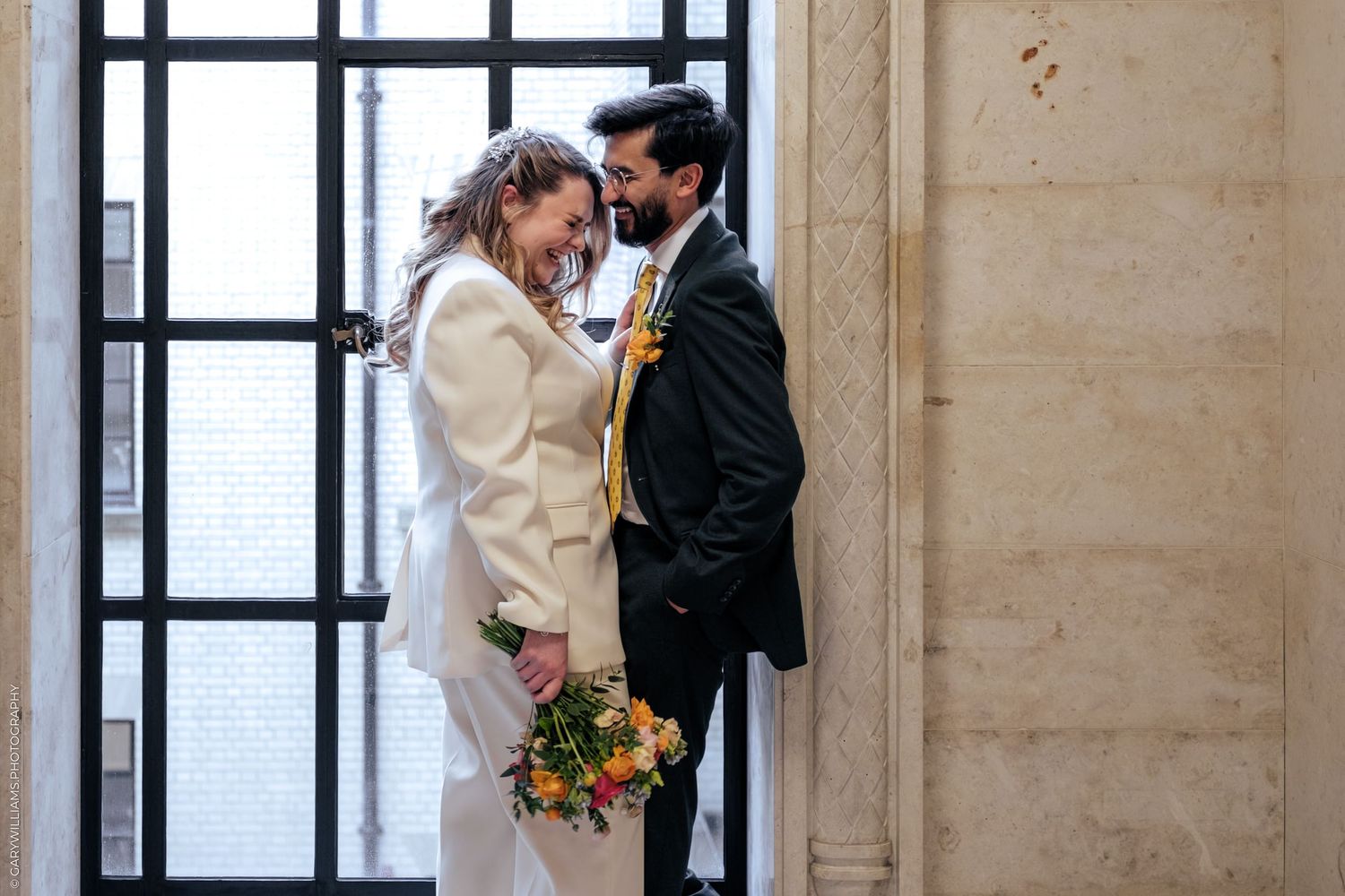 Bride and groom share a romantic moment in a sunlit hall, she holds a colorful bouquet, he wears a dark suit with yellow tie.