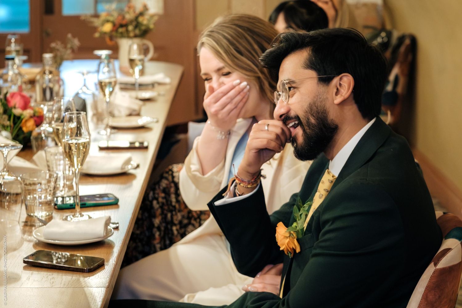 Couple laughing joyfully at a wedding reception dinner table, decorated with flowers and champagne glasses.