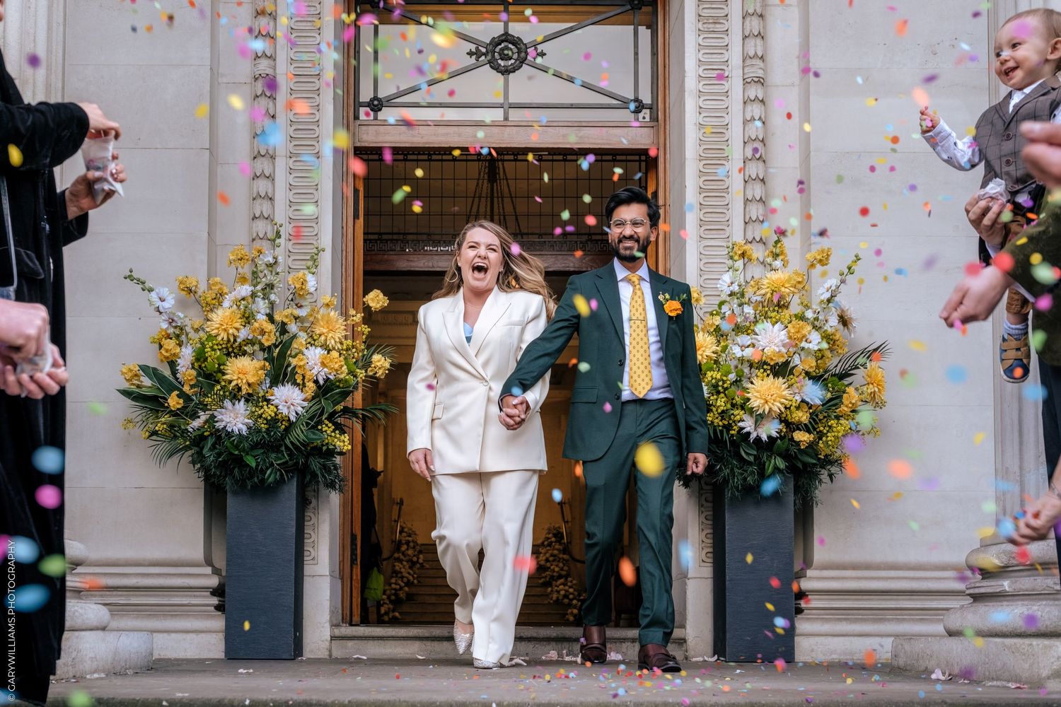 Newlyweds exit venue holding hands amid colorful confetti shower, bride in white suit, groom in green suit with yellow tie.