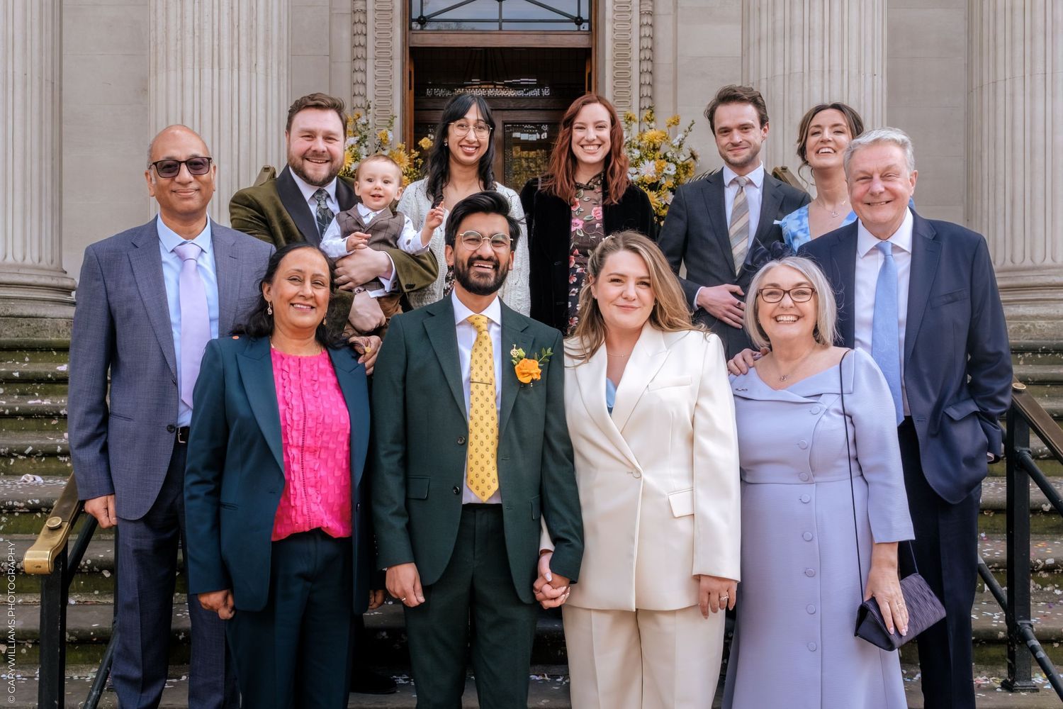 Wedding party group portrait on steps, bride in white suit and groom in green suit surrounded by smiling family members.