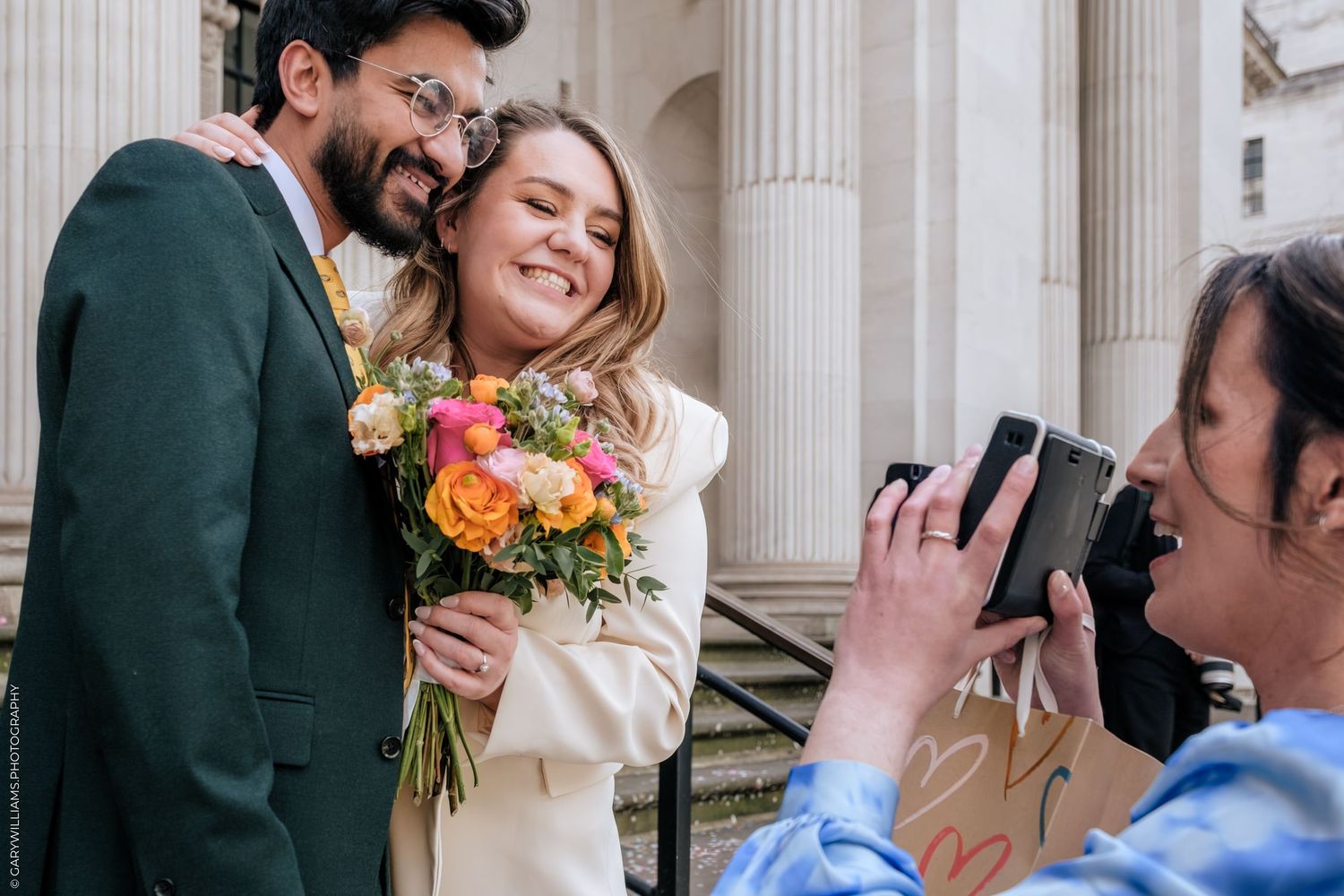Couple posing for wedding photos outside while photographer captures their joyful moment with colorful bouquet.