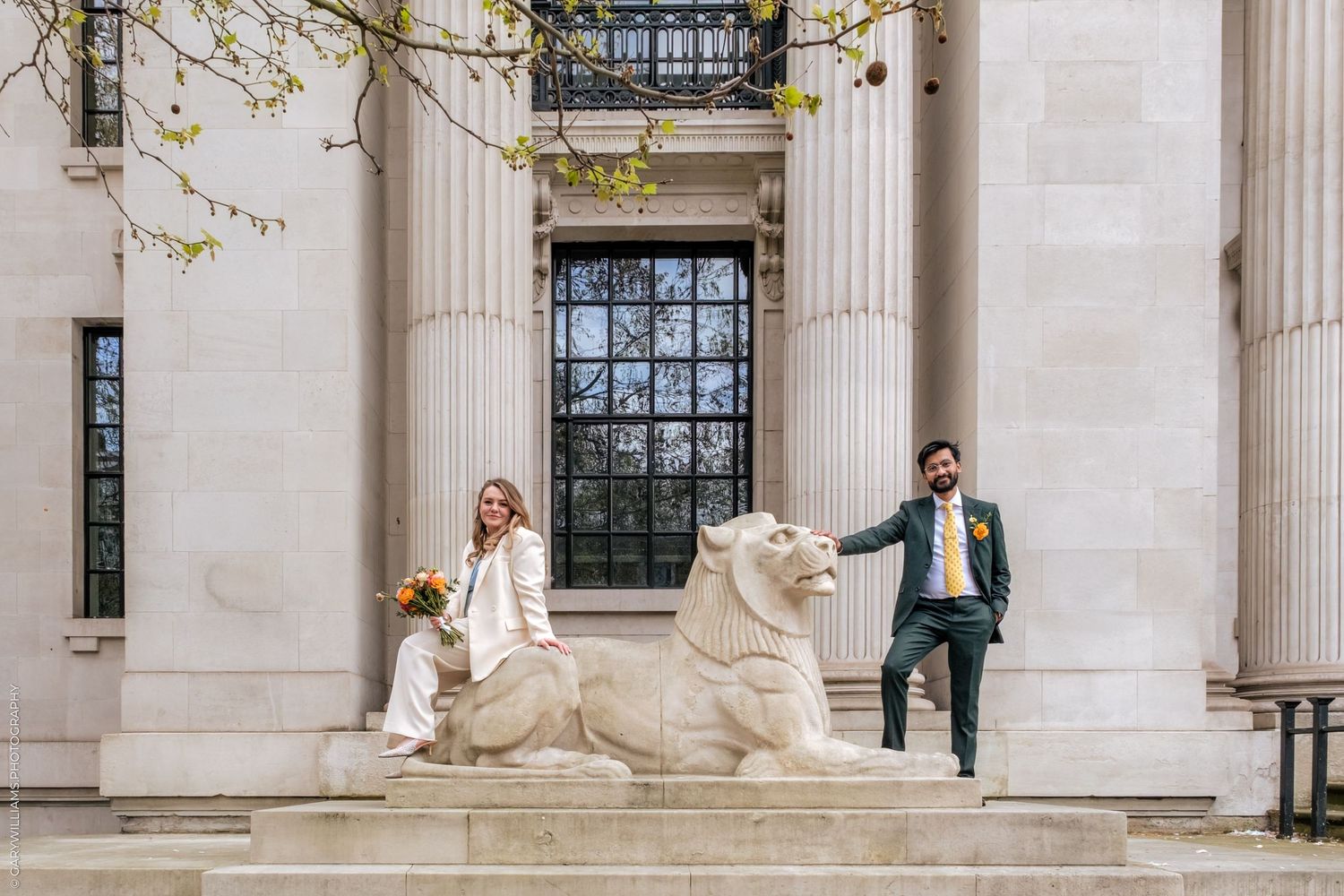 Bride and groom posing with a stone lion sculpture on steps of a neoclassical building with tall columns.