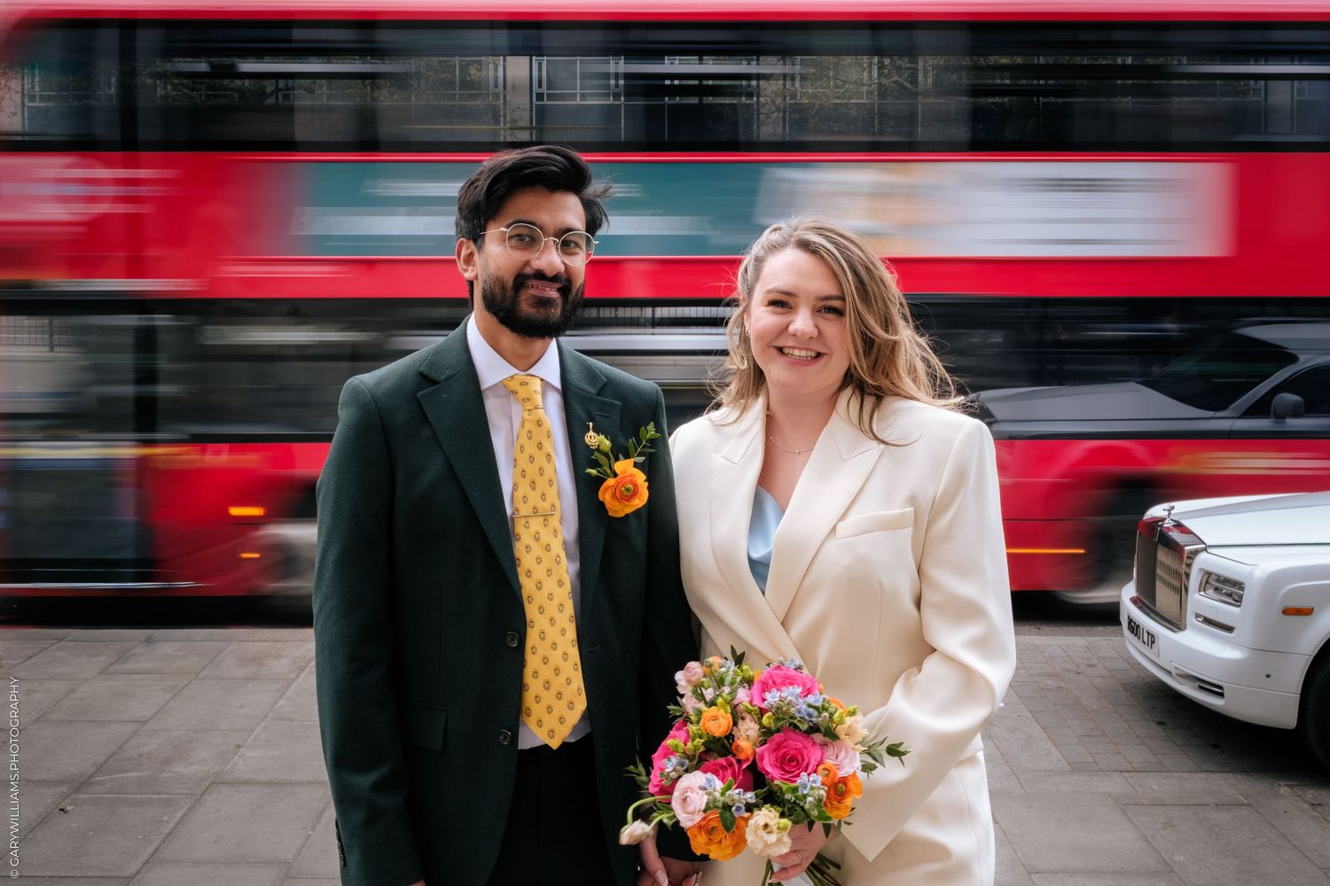 Bride and groom posing on London street with red double-decker bus and Rolls-Royce, bride holding colorful bouquet.