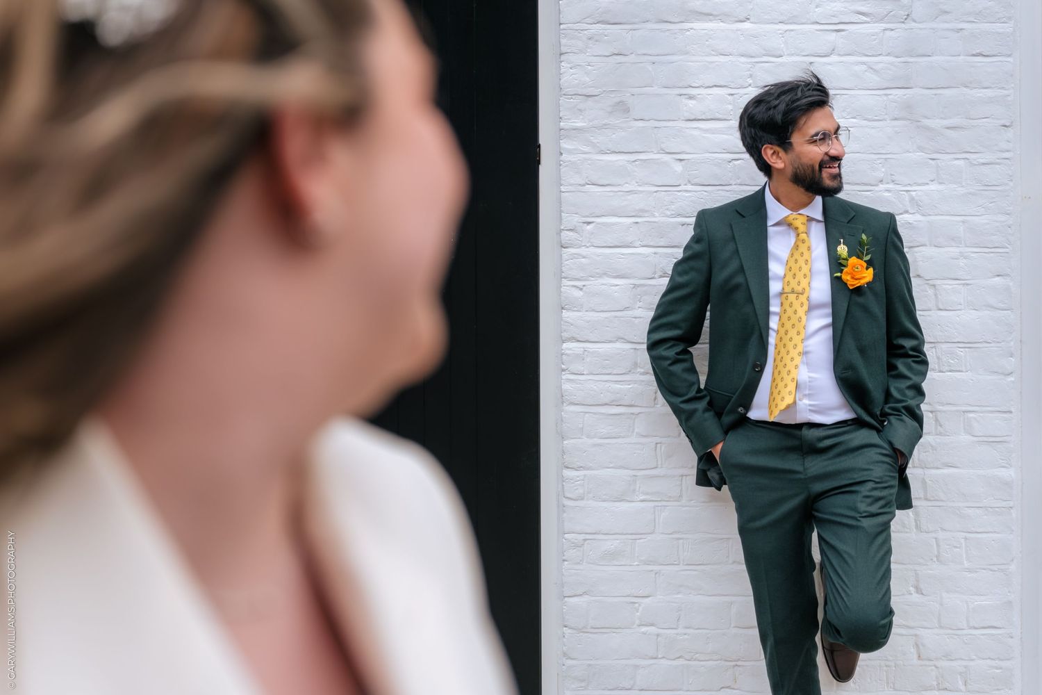 Groom in dark green suit with yellow tie and floral boutonniere leaning against white brick wall, bride in foreground.