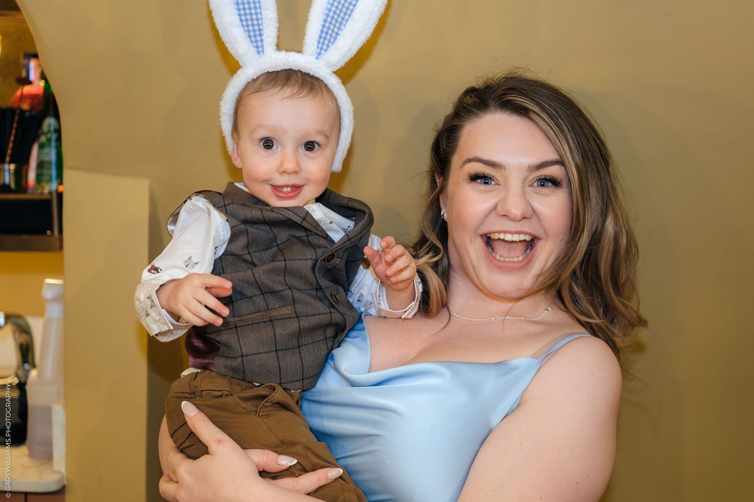 A joyful woman in a blue dress holds a baby boy wearing bunny ears at an Easter celebration.