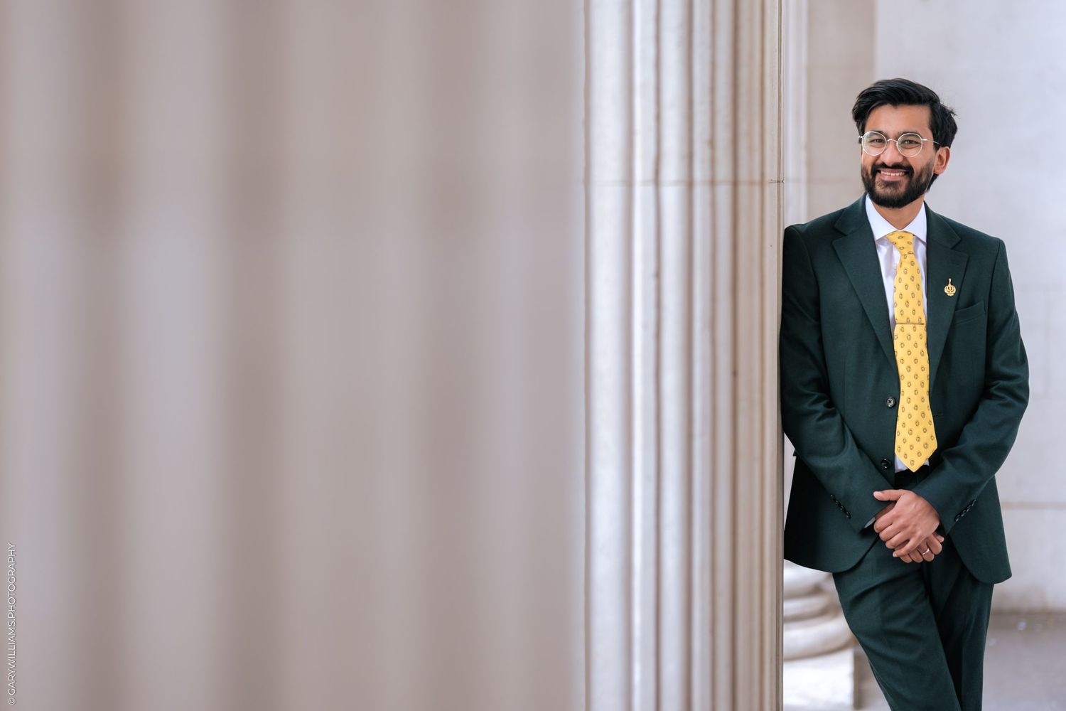 Man in dark green suit and yellow tie smiling while leaning against large stone columns.