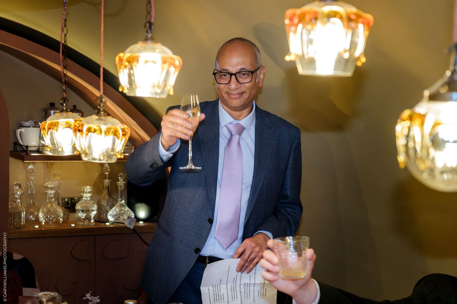 Elegant man in blue suit raising a champagne glass toast surrounded by vintage pendant lights in a restaurant.