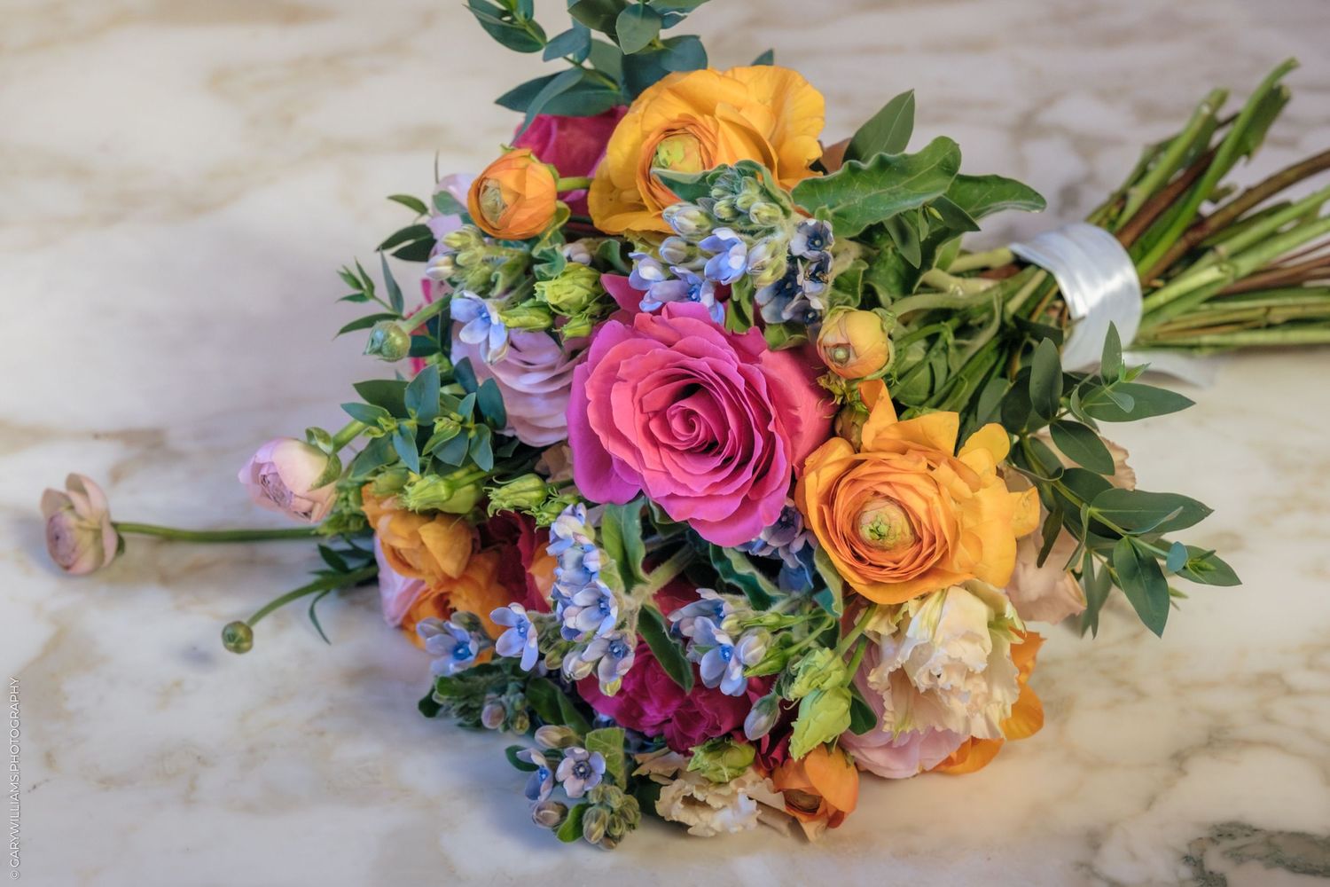 Colorful bridal bouquet with pink roses, orange ranunculus, and blue flowers on a marble surface.
