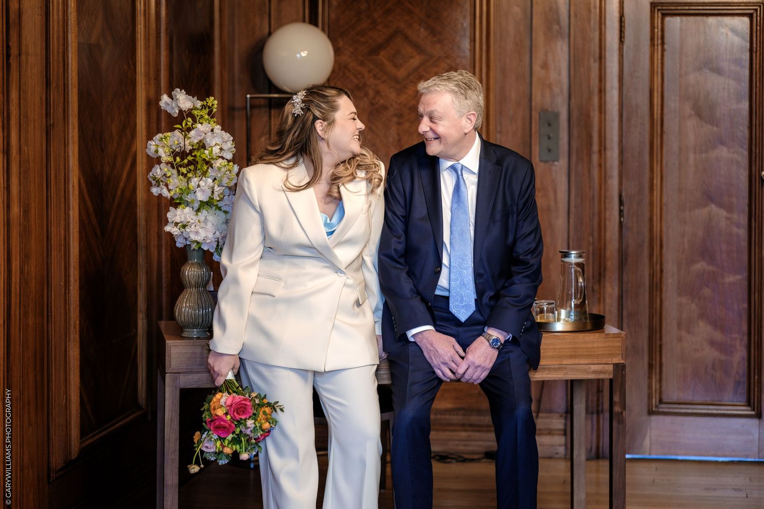 A bride in a white suit holds a colorful bouquet beside a man in a navy suit, sharing a smile in an elegant wood-paneled room.