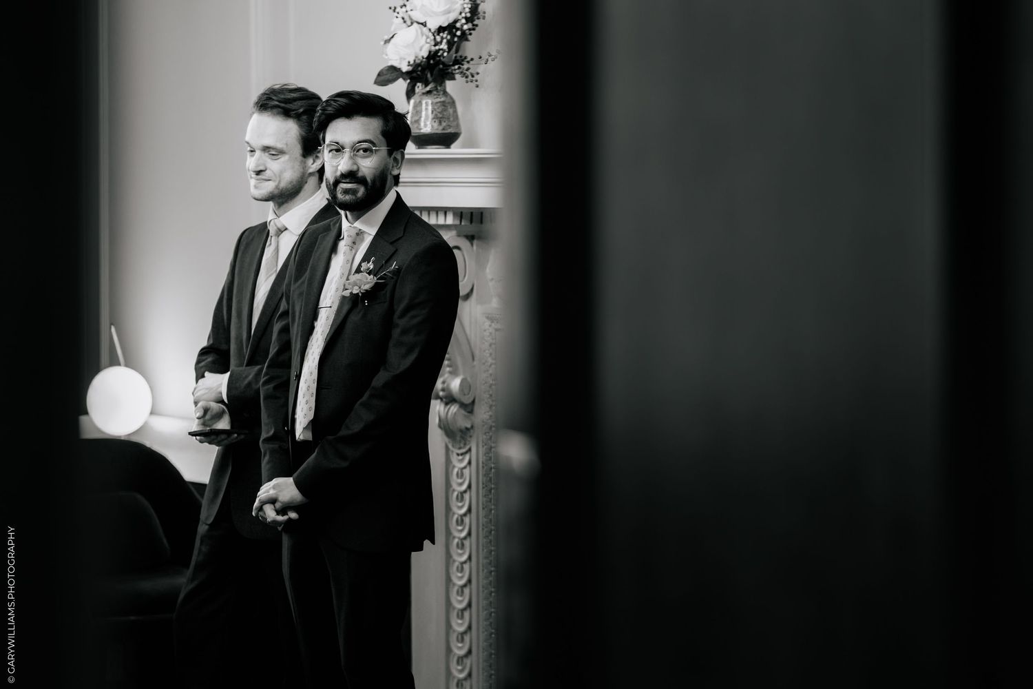 Two grooms in suits stand together during wedding ceremony in elegant venue, black and white photo.