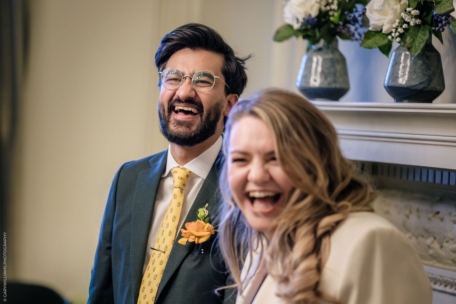 Joyful couple laughing together at their wedding ceremony, groom in navy suit with yellow tie and orange boutonniere.