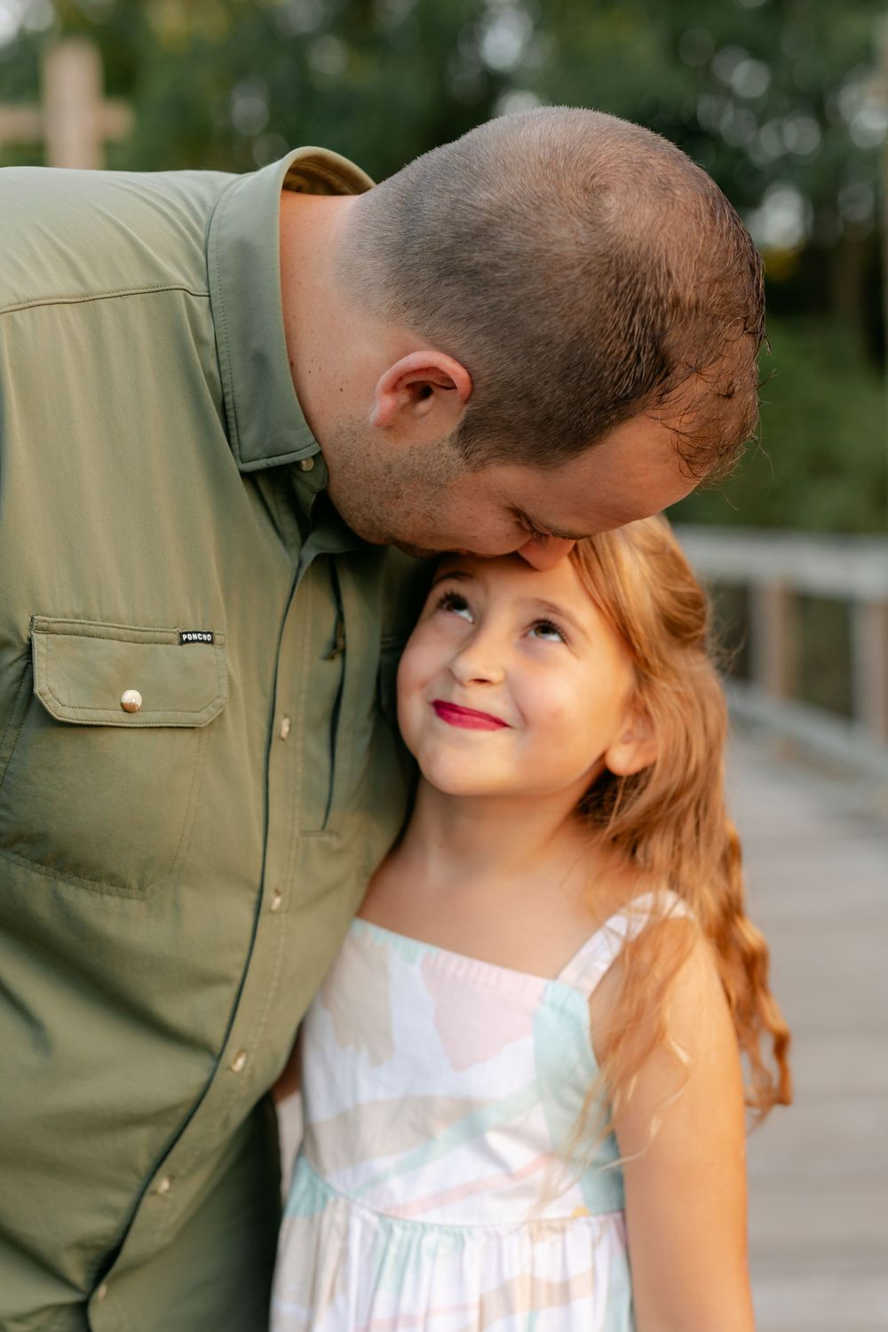 A loving father kisses his smiling young daughters forehead on a wooden bridge at golden hour.