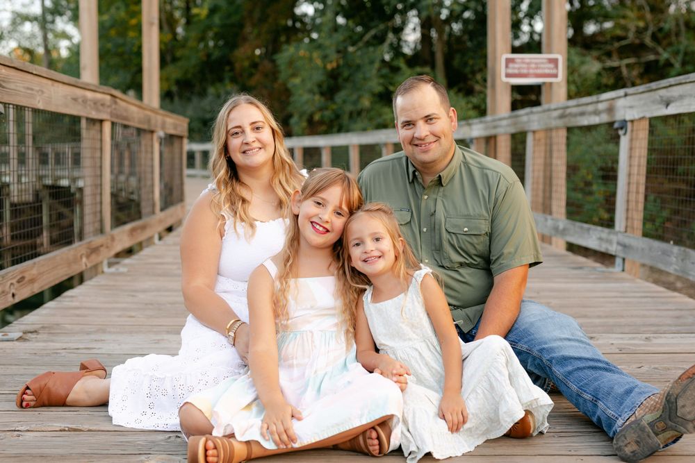 A happy family of four sits together smiling on a wooden bridge surrounded by lush greenery.