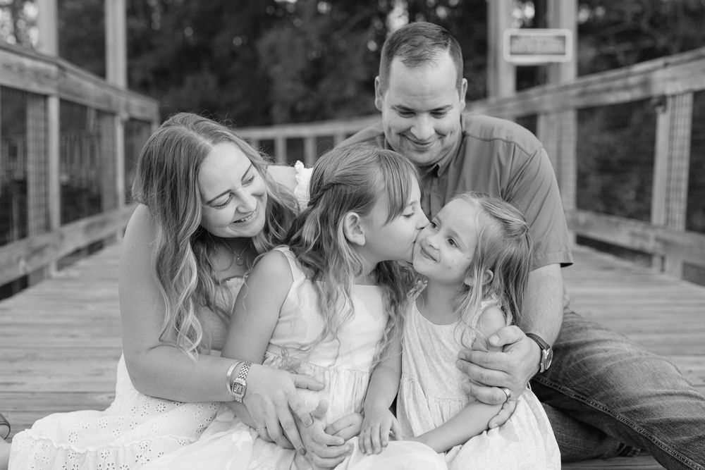 Black and white family photo of parents and two daughters sharing a sweet moment on a wooden bridge.