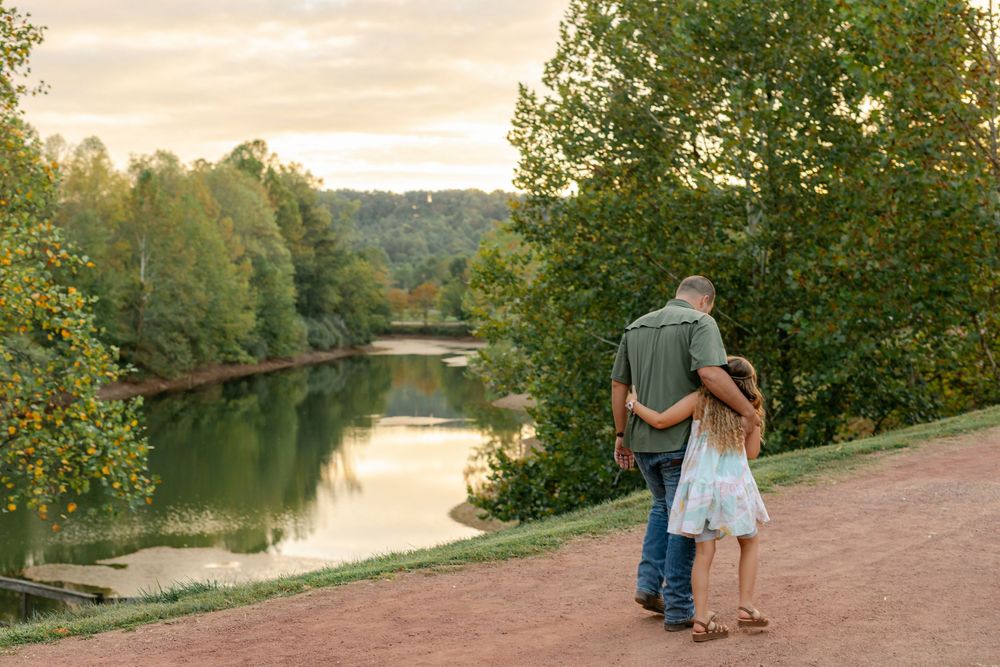 A couple shares a romantic embrace overlooking a serene lake surrounded by lush green trees.