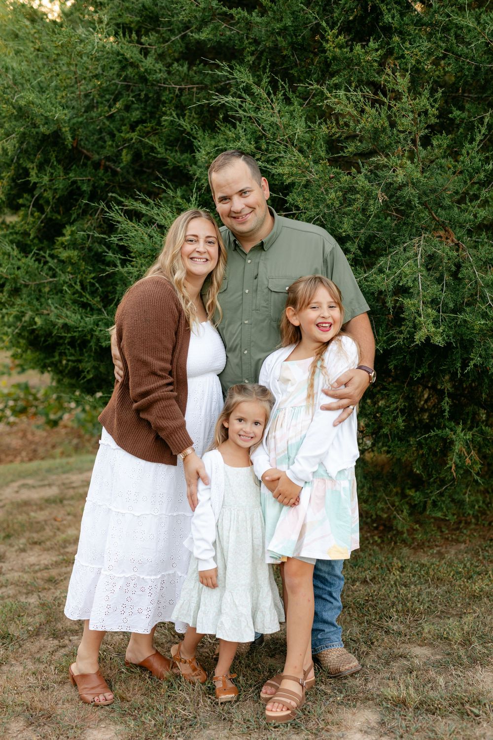 A smiling family of four poses together in front of tall green evergreen trees outdoors.