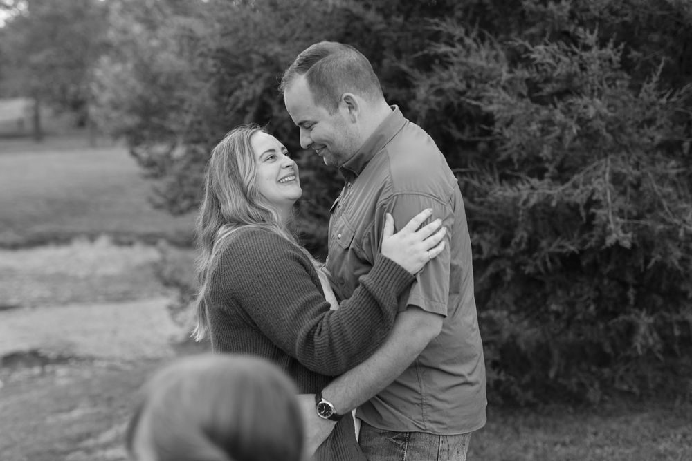 Black and white photo of a smiling couple gazing lovingly at each other outdoors near trees.