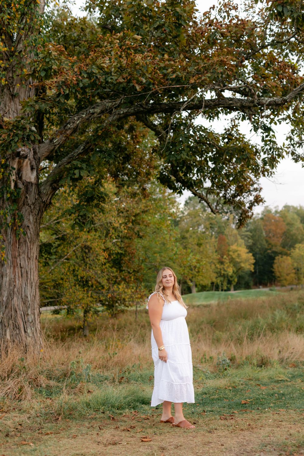 A blonde woman in a white sundress stands gracefully beneath a large autumn tree in an open field.