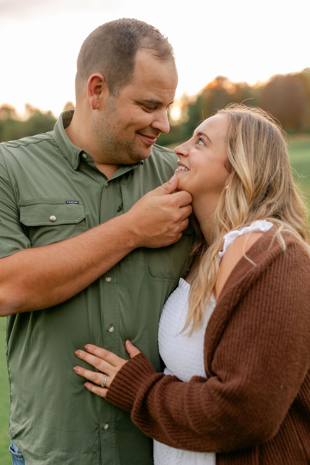 A loving couple smiles at each other in a golden sunset field, woman wearing an engagement ring.