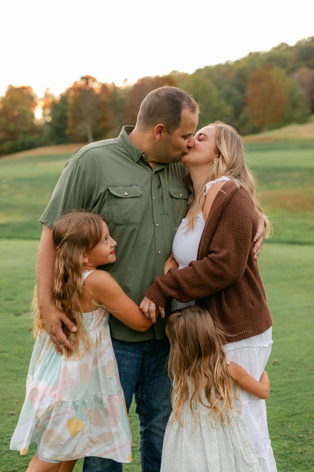 A family of four shares a sweet kiss moment together on a green lawn at golden sunset.