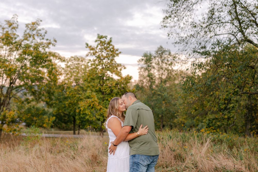 Couple sharing a romantic kiss in a golden field with lush green trees and cloudy sky backdrop.