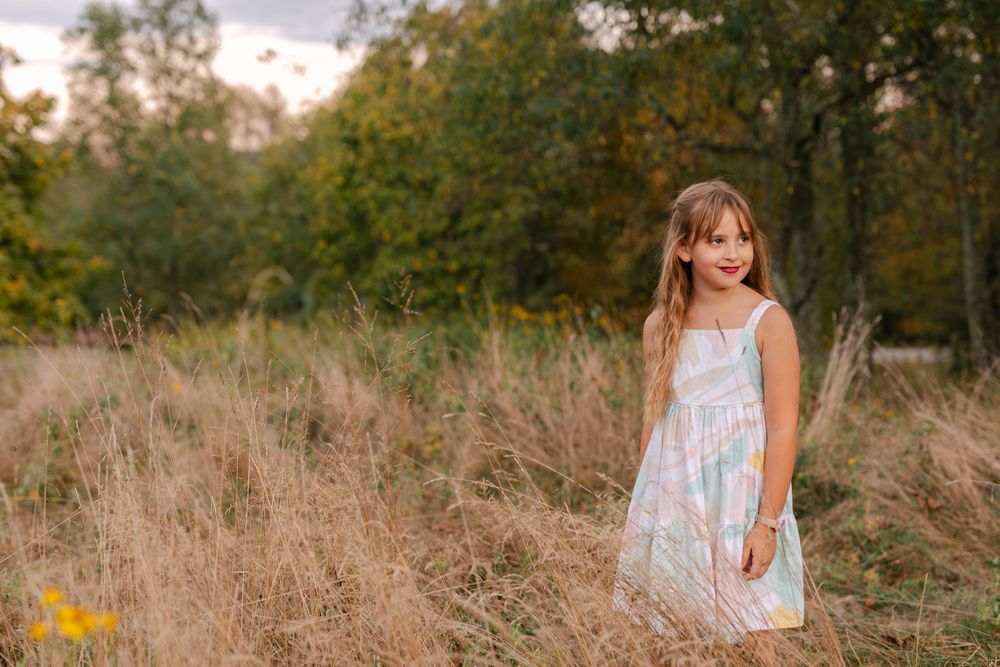 Smiling young girl in a pastel floral dress posing in a golden autumn meadow with trees behind.