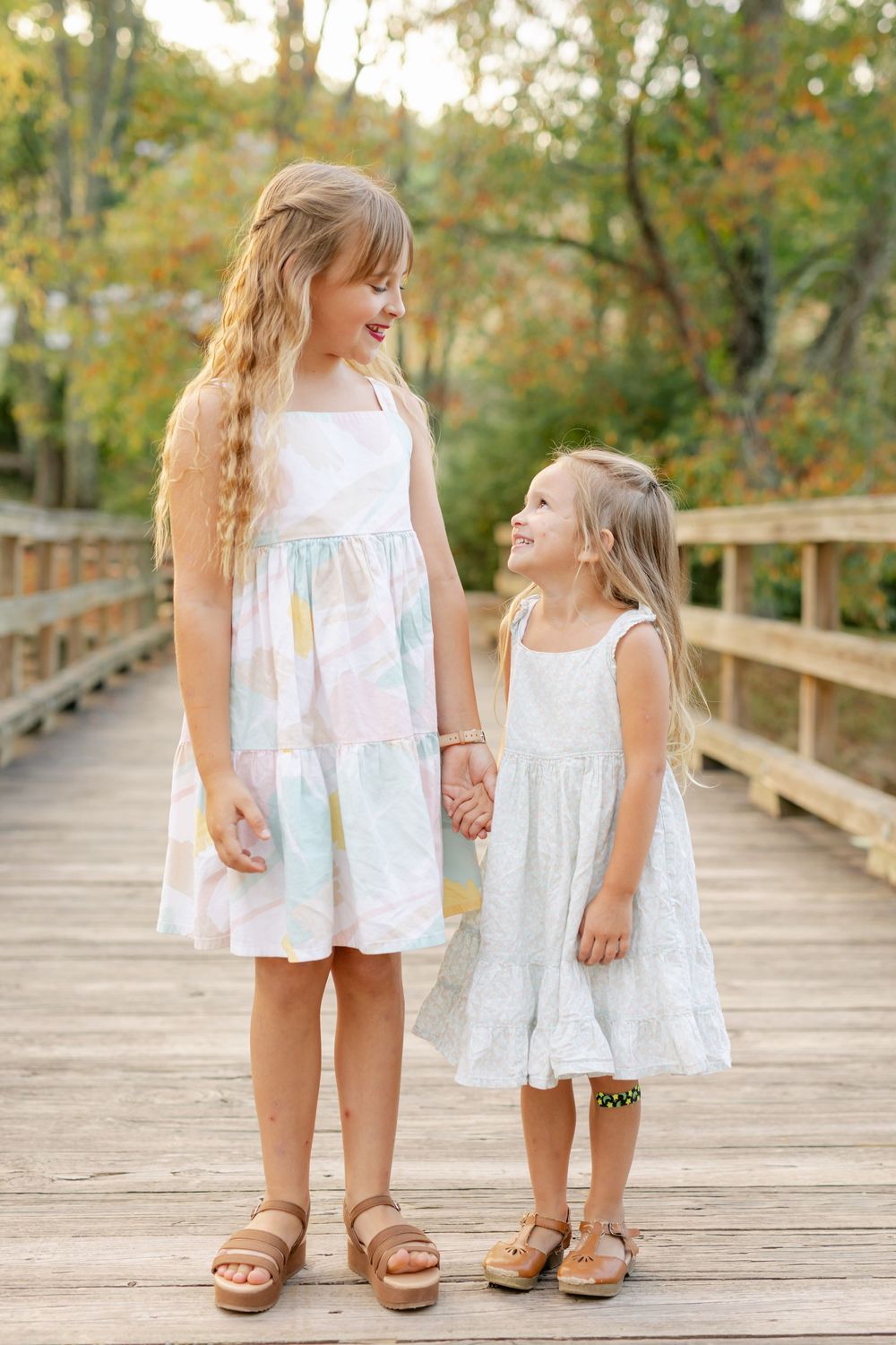 Two young sisters in white and pastel dresses hold hands and laugh on a wooden bridge.