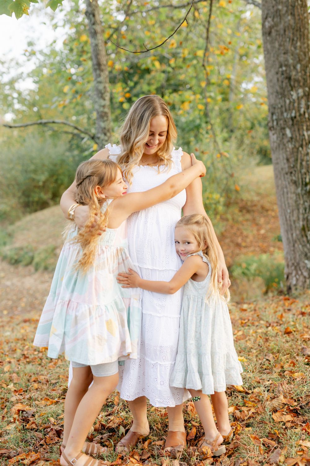 Pregnant mother in white dress shares a tender embrace with her two young daughters among autumn leaves.