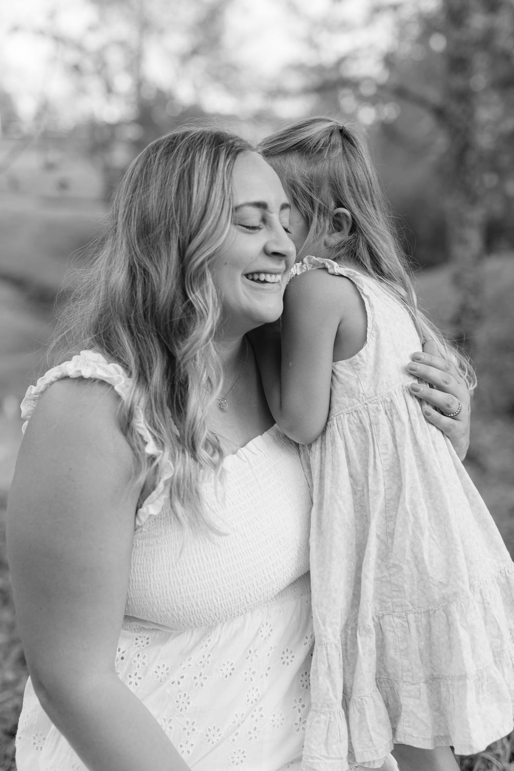 Black and white photo of a little girl hugging and kissing her smiling mother's cheek outdoors.