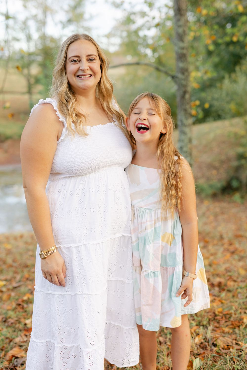 A smiling blonde mother in a white dress poses with her laughing daughter in a colorful fall dress.