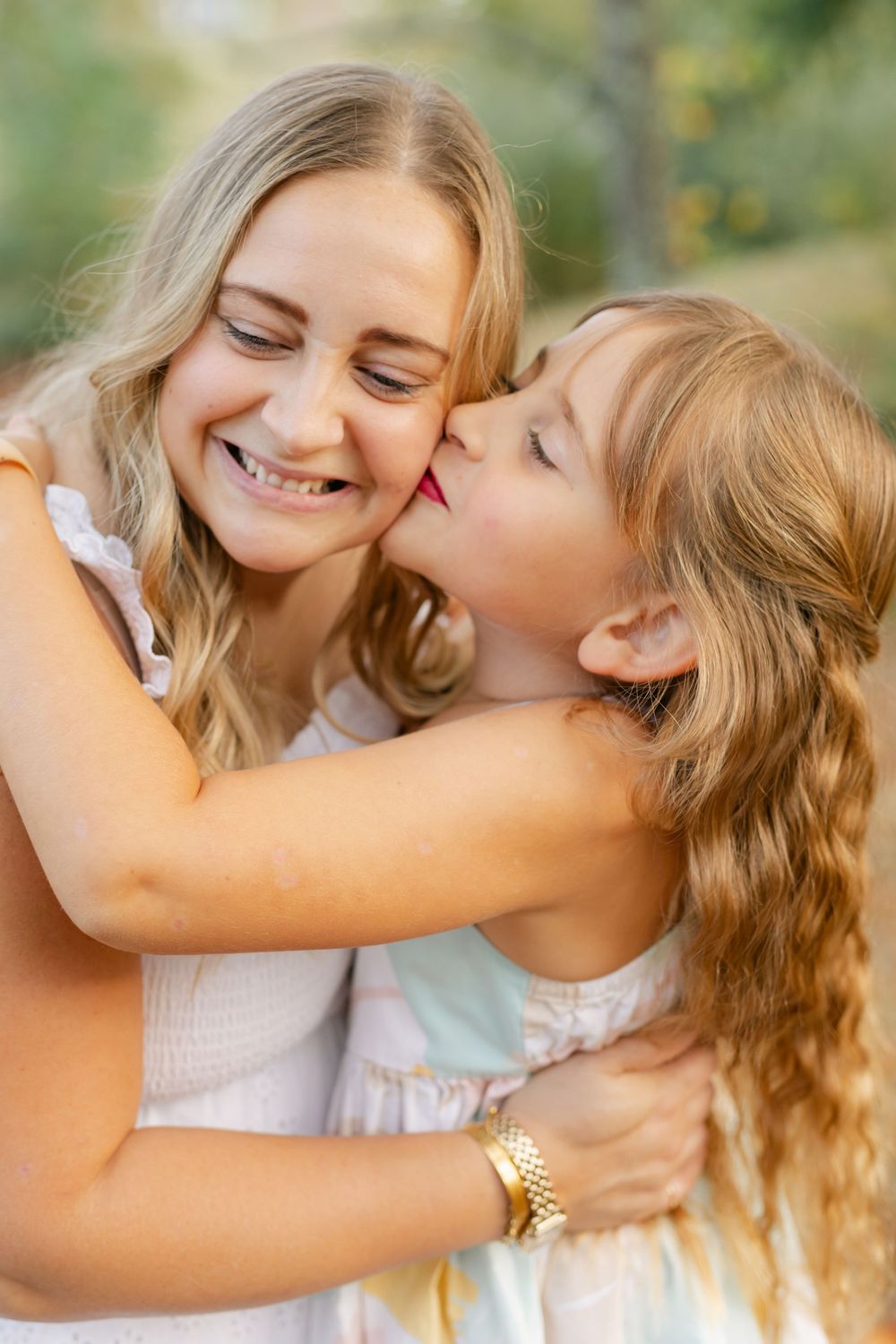 A young girl in a light dress gives her smiling blonde mother a kiss on the cheek outdoors.