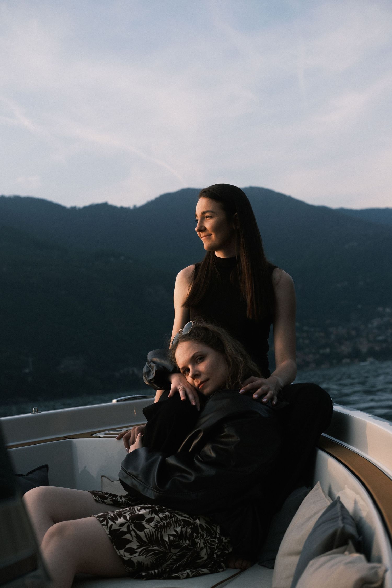 Two young women relaxing on a luxury boat with mountain scenery and cloudy sky in the background.
