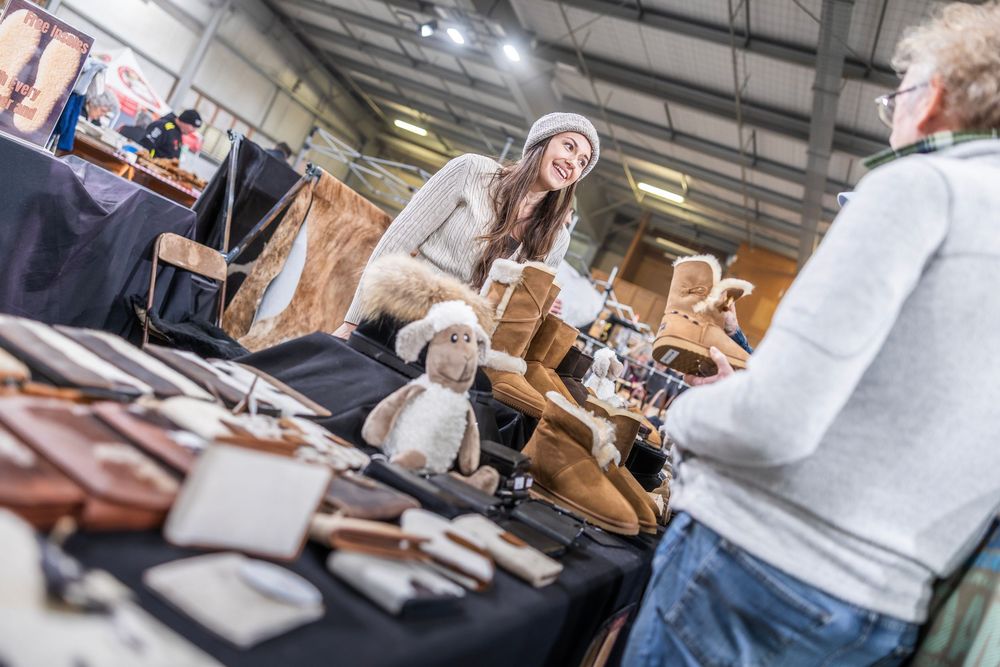 Woman smiling at a market stall selling UGG boots, leather goods, and a sheep stuffed animal inside an indoor venue.