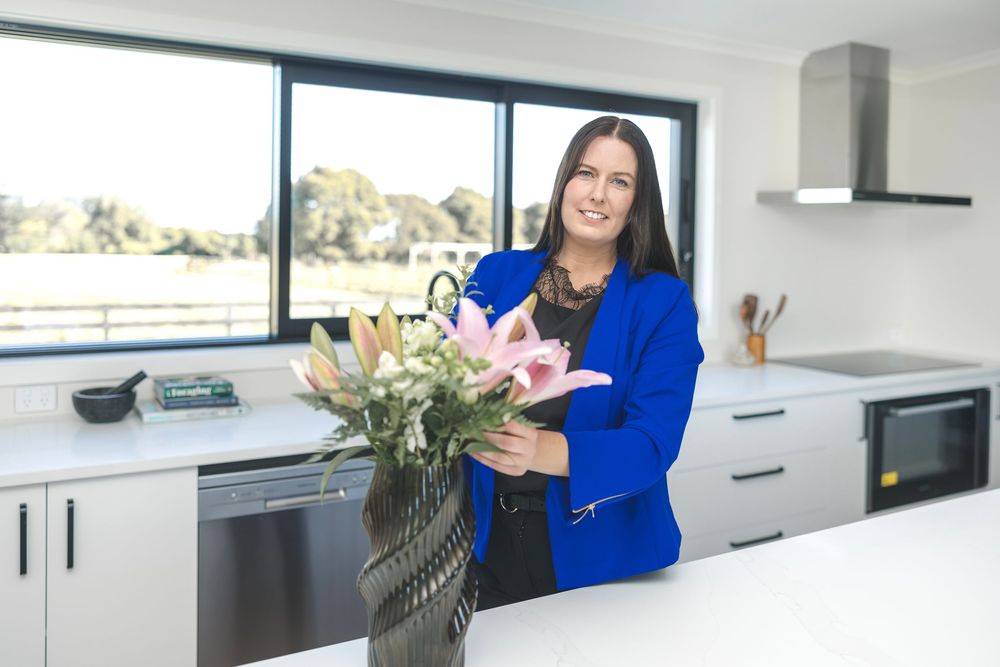 Woman in blue blazer arranging pink lilies in a vase on a modern white kitchen island.