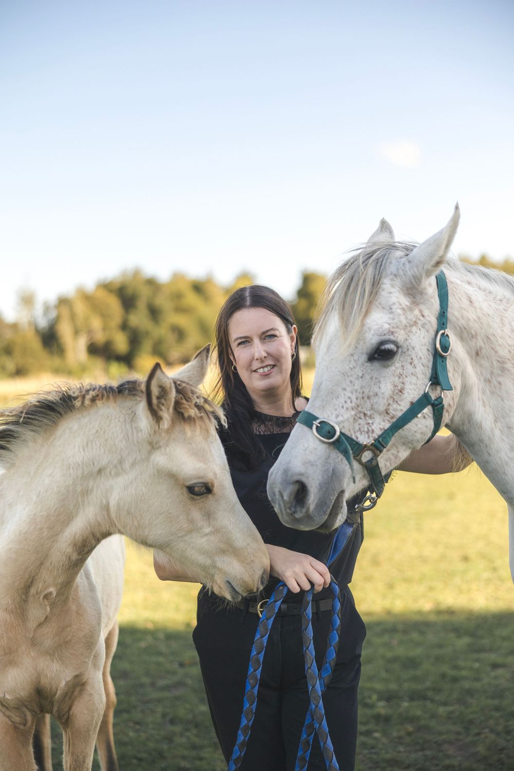 Woman smiling while holding a white dapple horse and a young foal in a sunny green pasture.