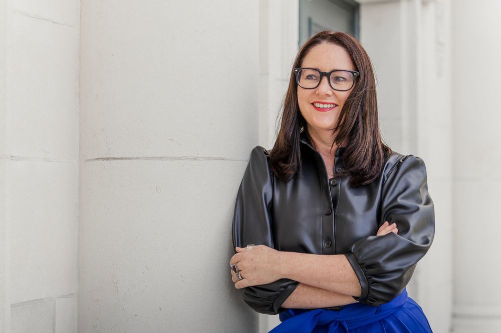 Professional woman in glasses and leather jacket smiling with arms crossed against a white wall.