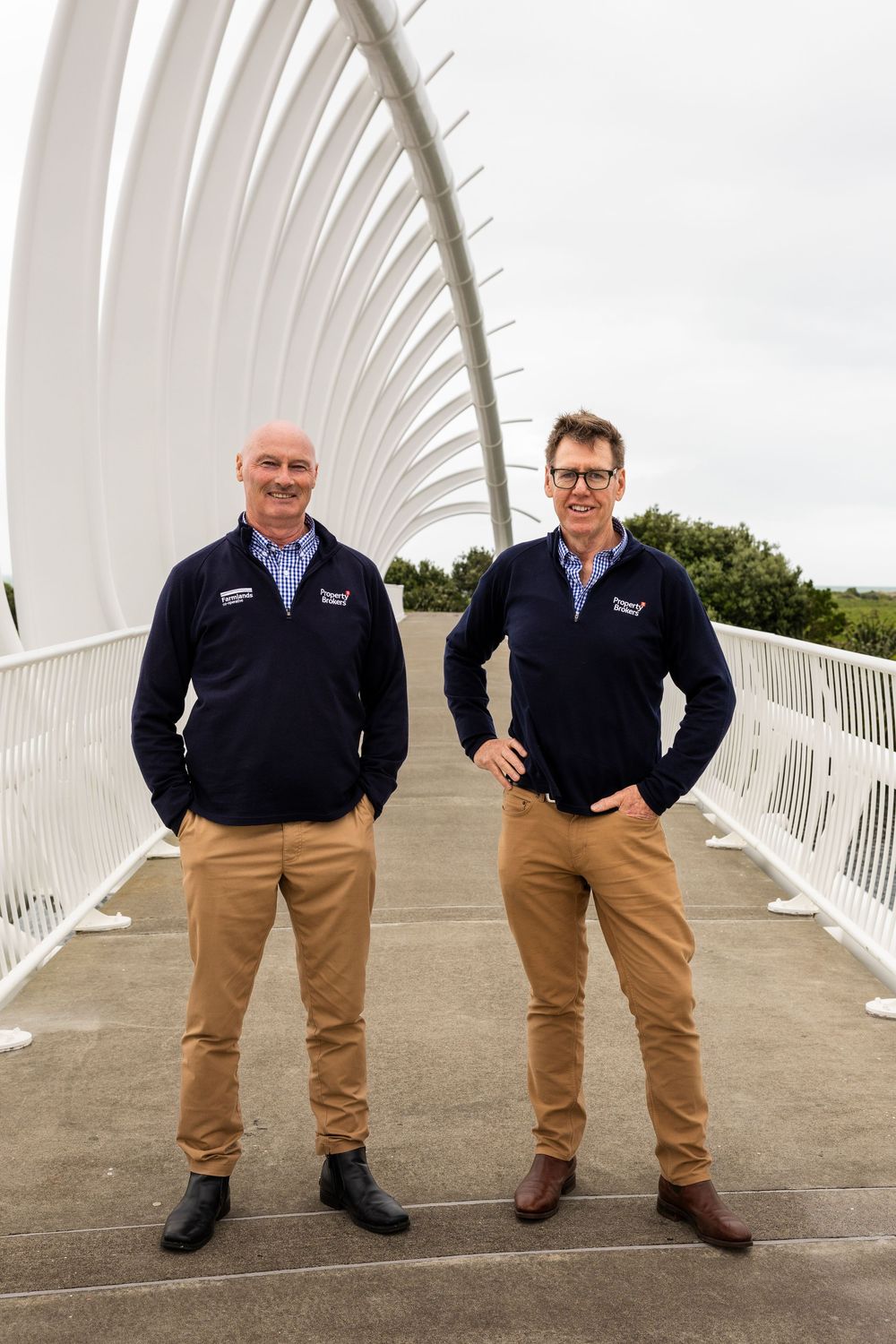 Two men in matching navy fleeces and khaki pants stand on a modern white arched bridge outdoors.