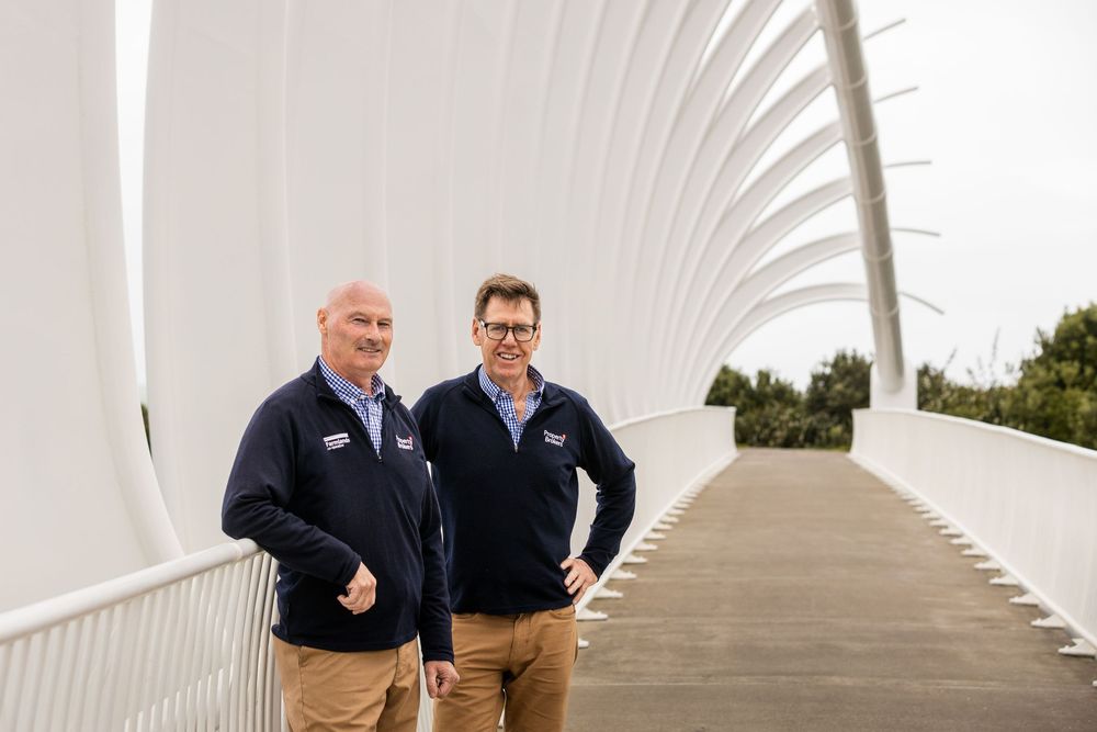 Two men in navy fleeces stand on a white arched pedestrian bridge walkway.