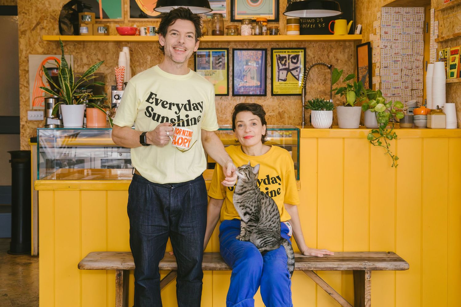 Two people in yellow Everyday Sunshine shirts sit outside a colorful cafe with RUDE signage and a QR code on the window.