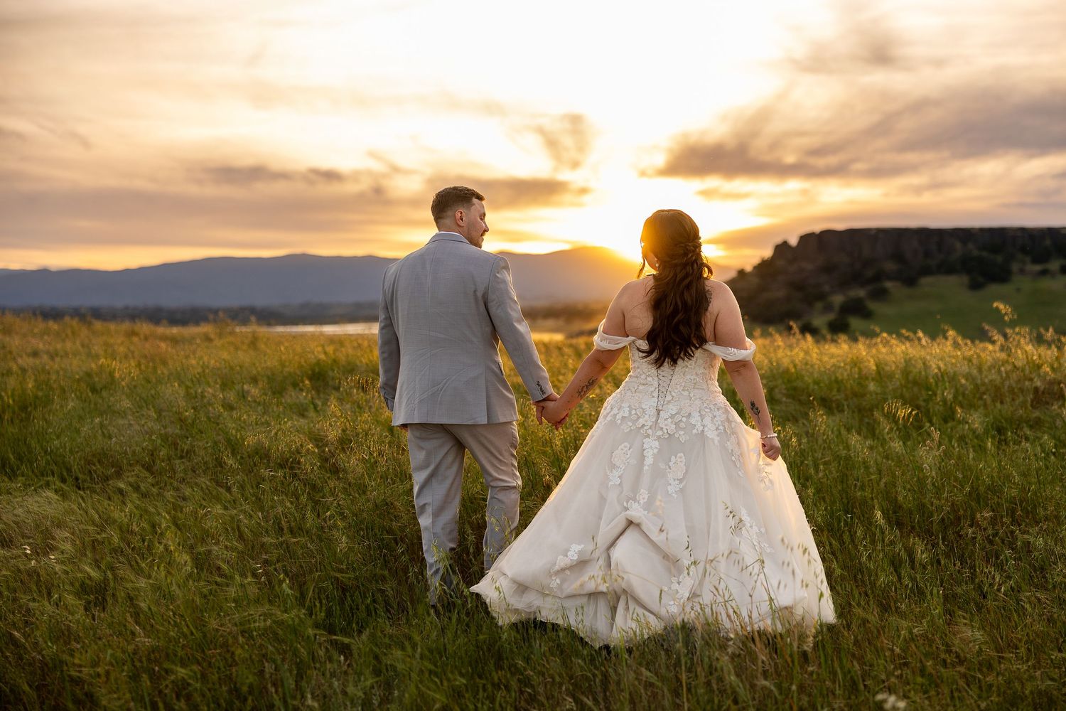 Bride and groom walk hand-in-hand through a golden field at sunset, she in lace gown, he in gray suit.