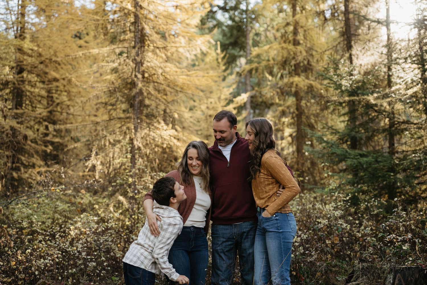 Family of four laughing together in an autumn forest with golden trees during a fall outdoor photo session.