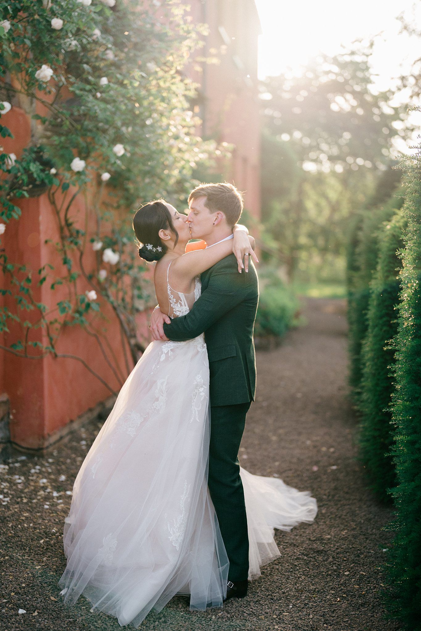A bride and groom share a romantic kiss in a sunlit garden, surrounded by lush greenery and climbing roses.