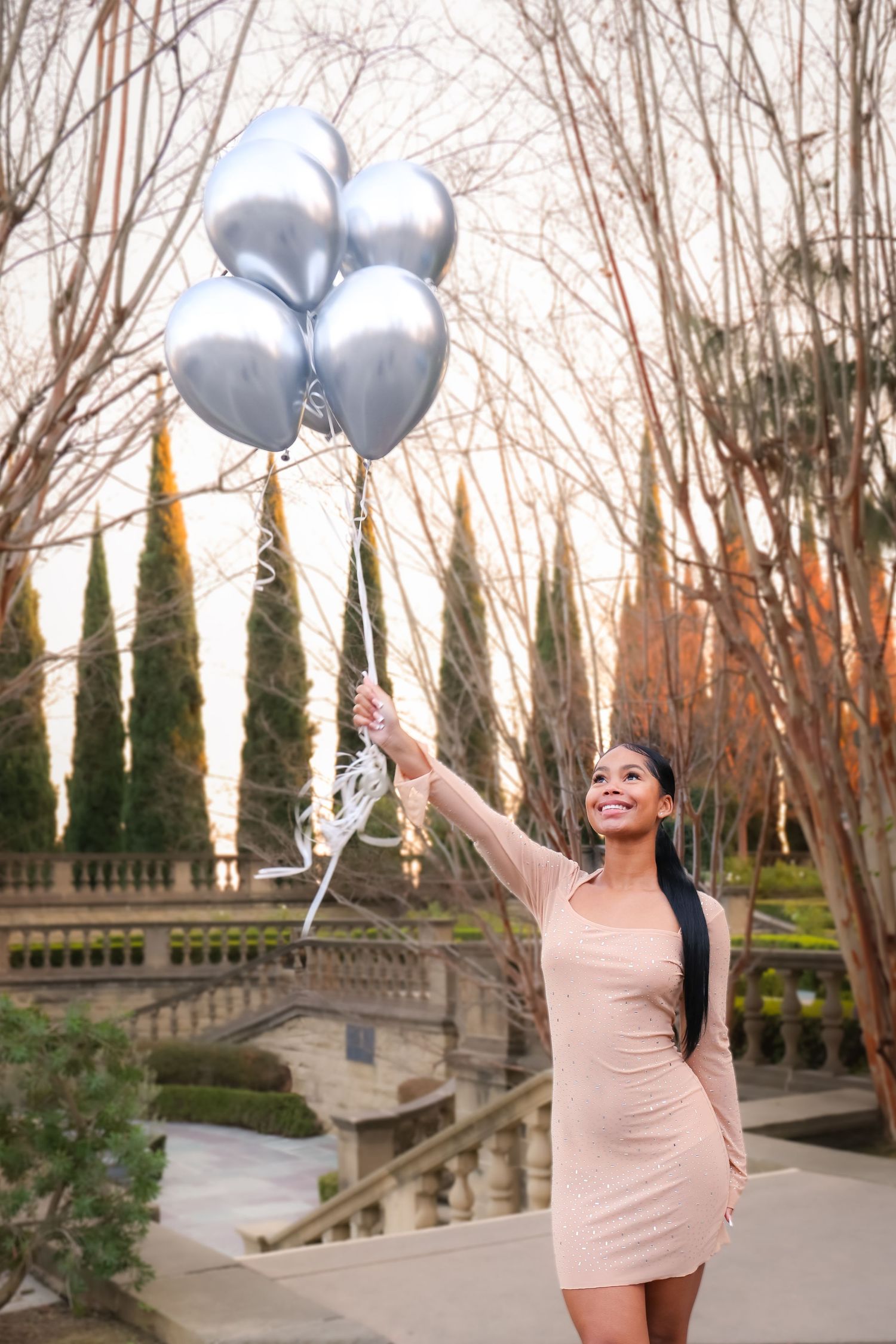 Young woman in a pink mini dress joyfully holding silver metallic balloons in a park at dusk.