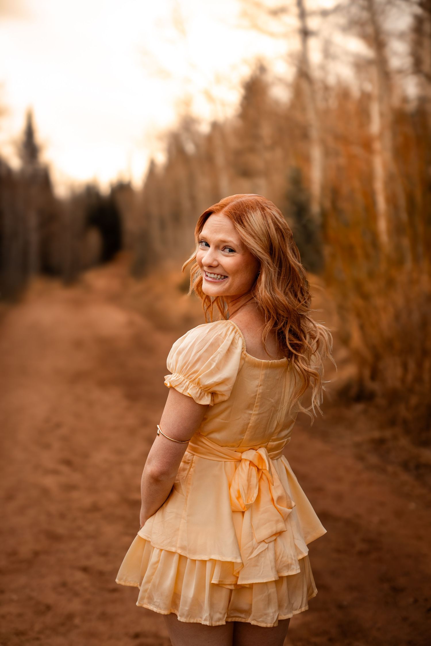 Smiling redhead woman in yellow ruffled dress on autumn dirt path surrounded by golden trees.