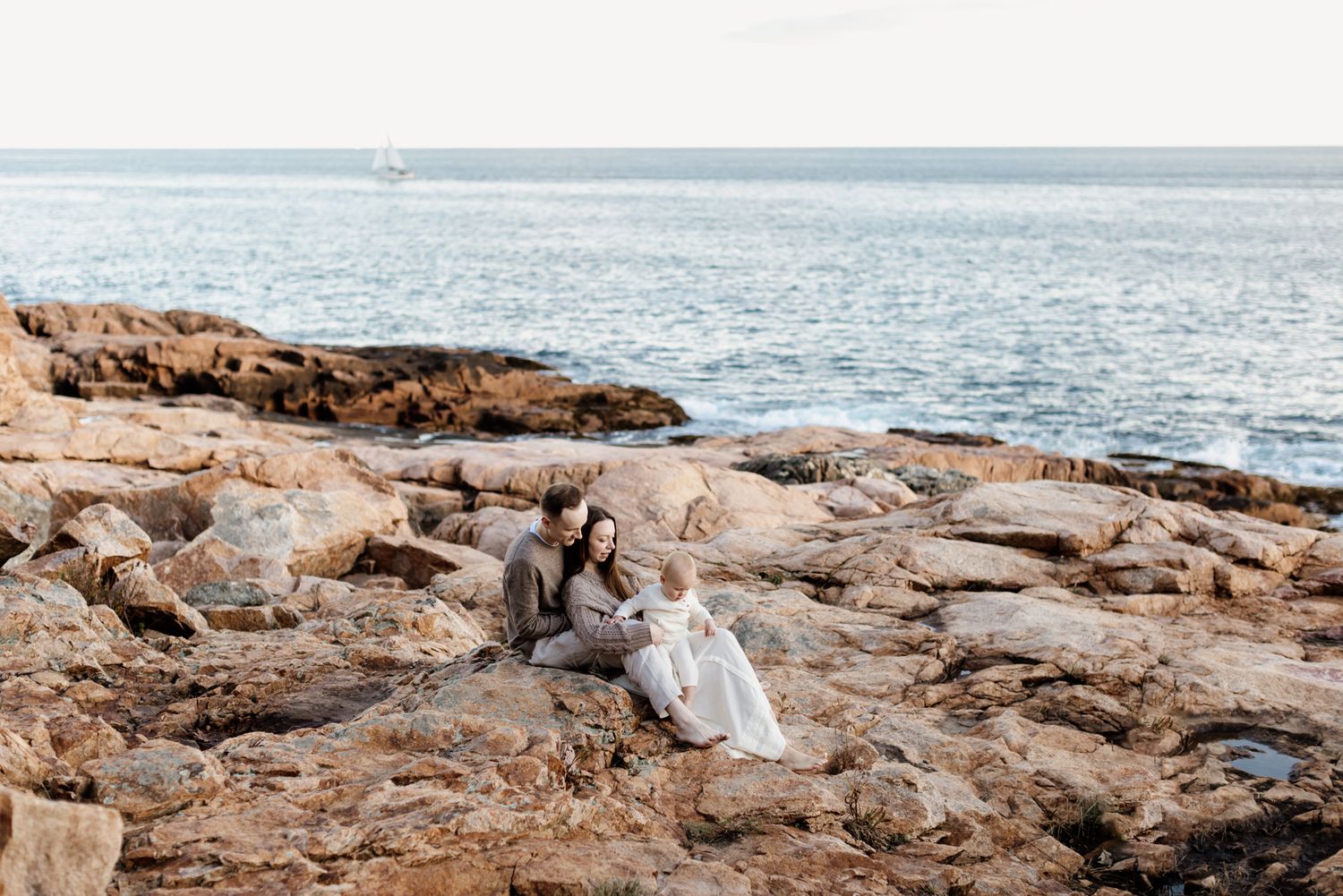 Family in neutral clothes sits on large coastal rocks with blue ocean water in background for family photos in Gloucester North Shore MA.